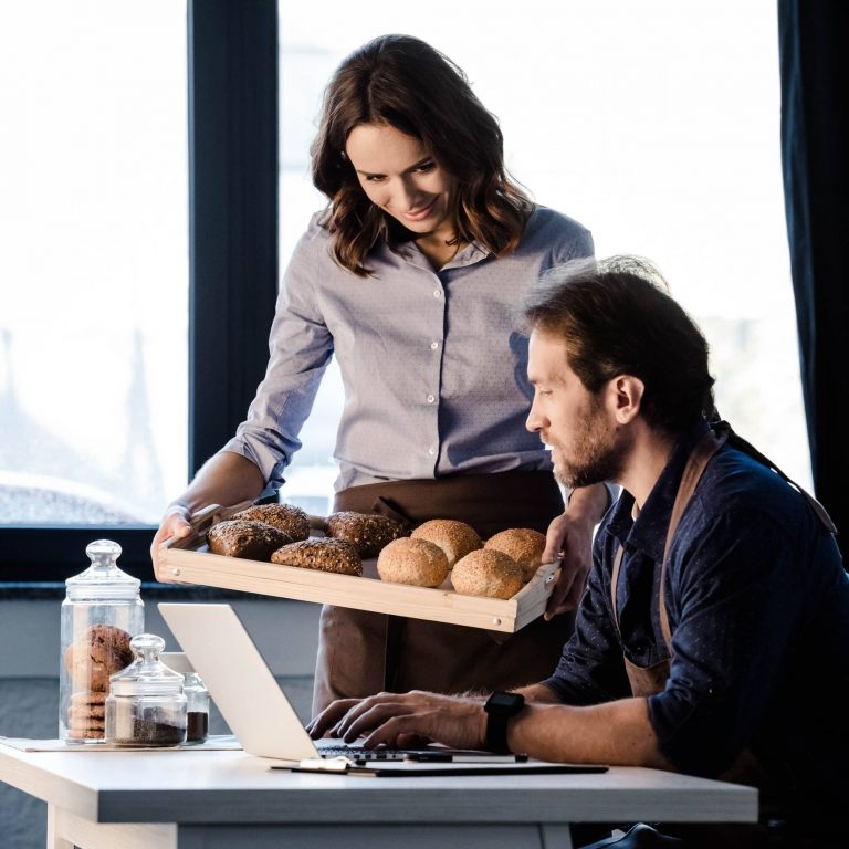 Male and female bakers examining pastries and using laptop in bakery